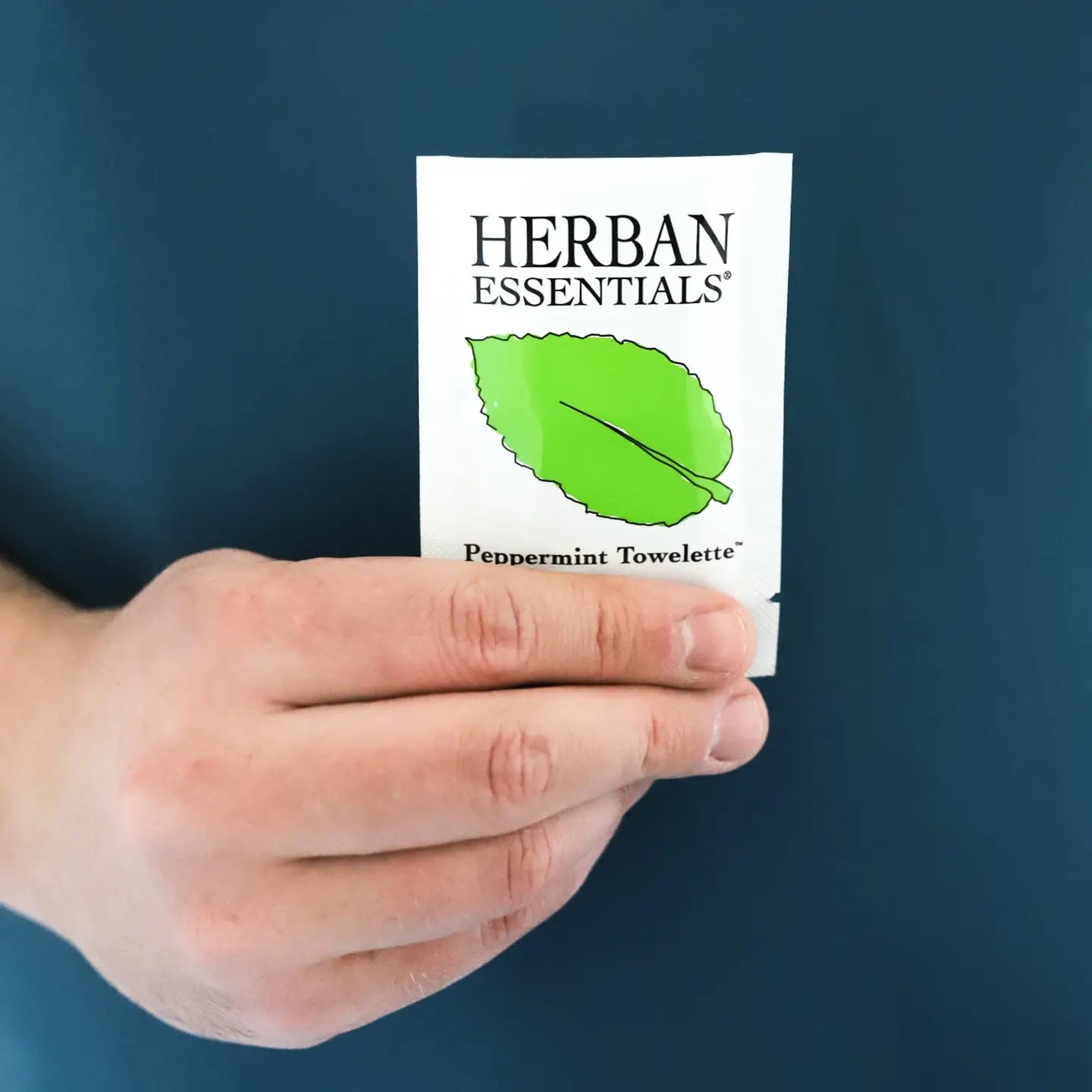 Hand holding a Herban Essentials peppermint towelette against a blue background