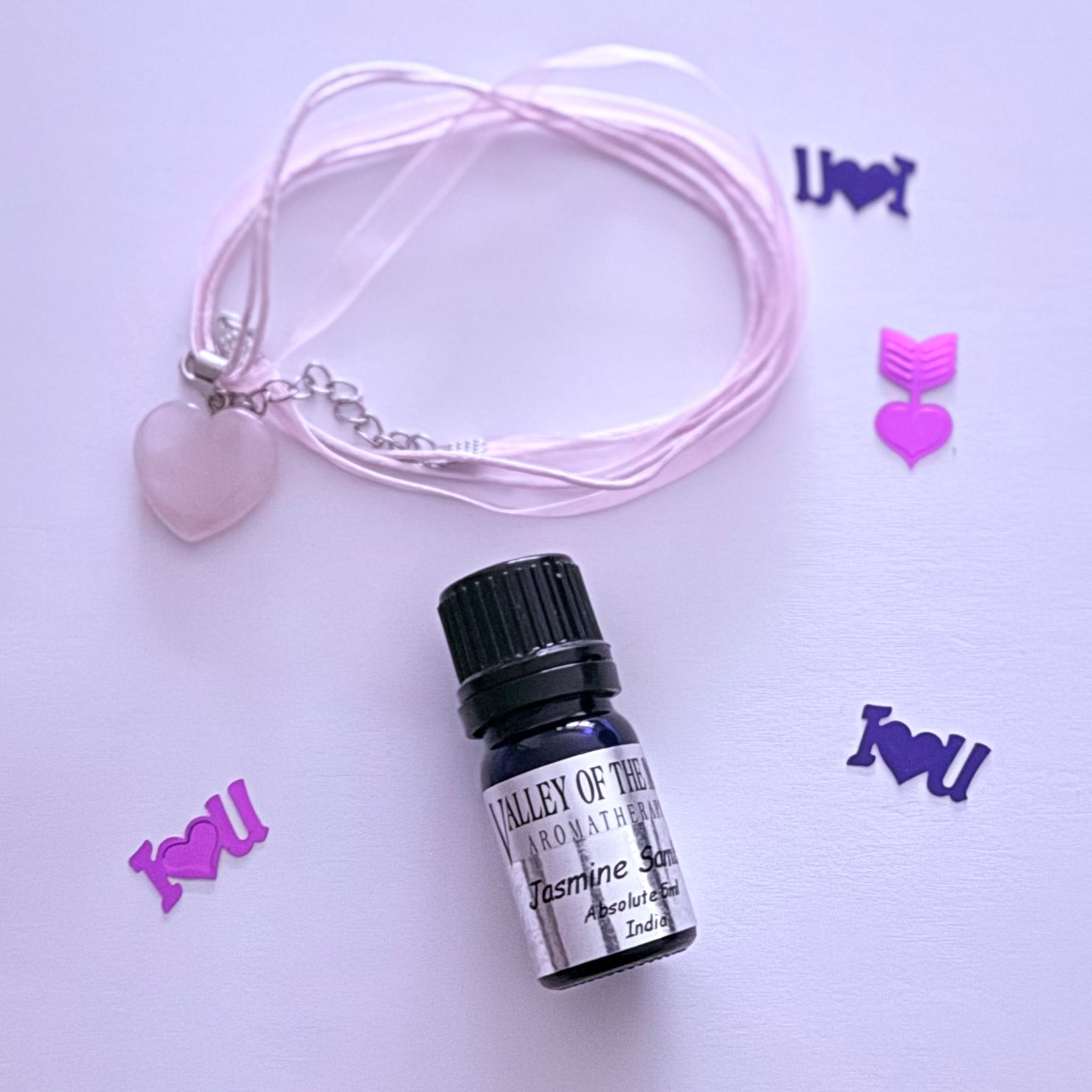 Small bottle of Jasmine Sambac essential oil with pink necklace and heart charm on a light purple background.