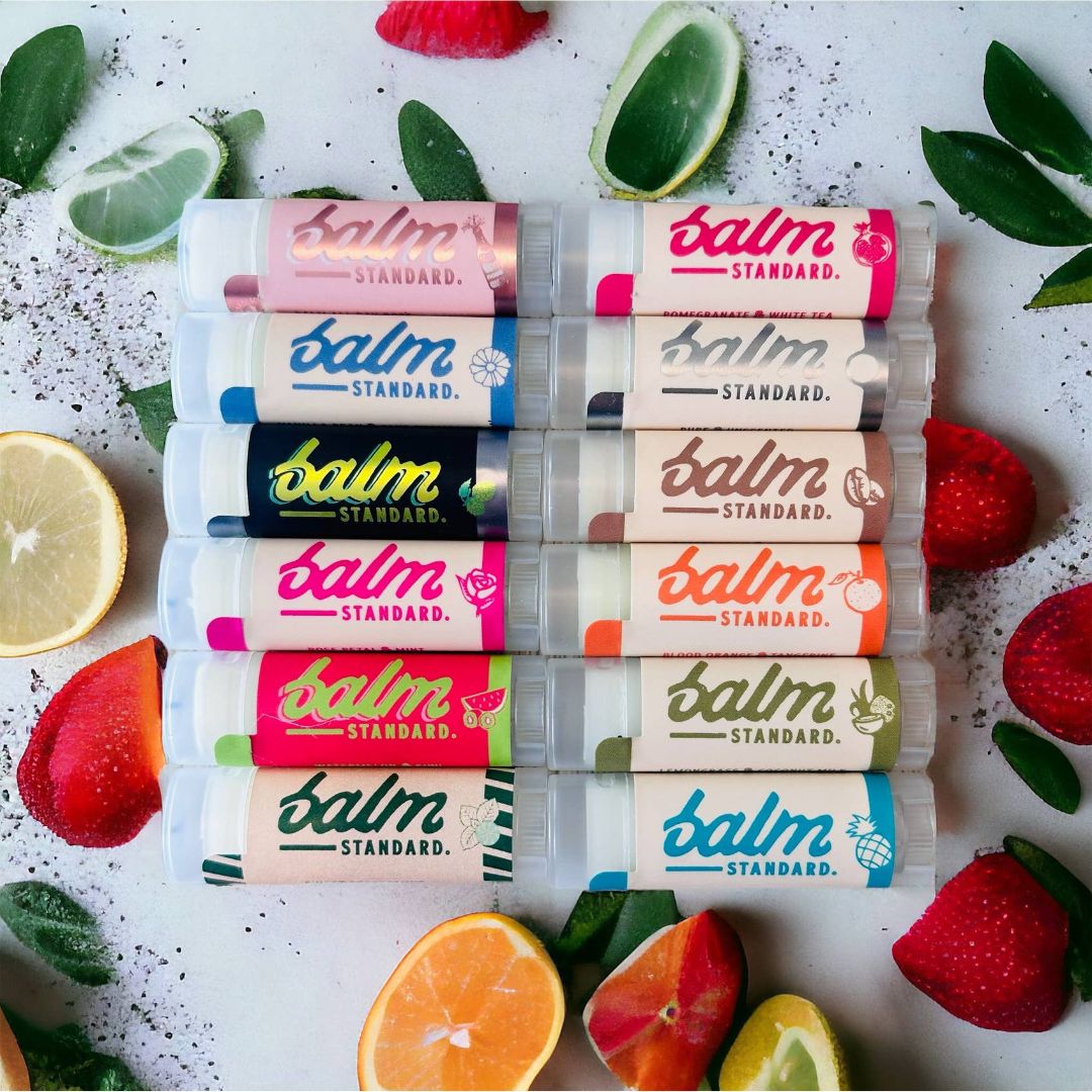 Collection of 'balm Standard' lip balms with colorful packaging on a white background with fruits and leaves.